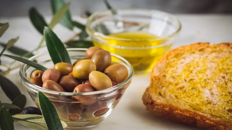 Pieces of bread being dipped in a bowl of olive oil next to bowls of olives