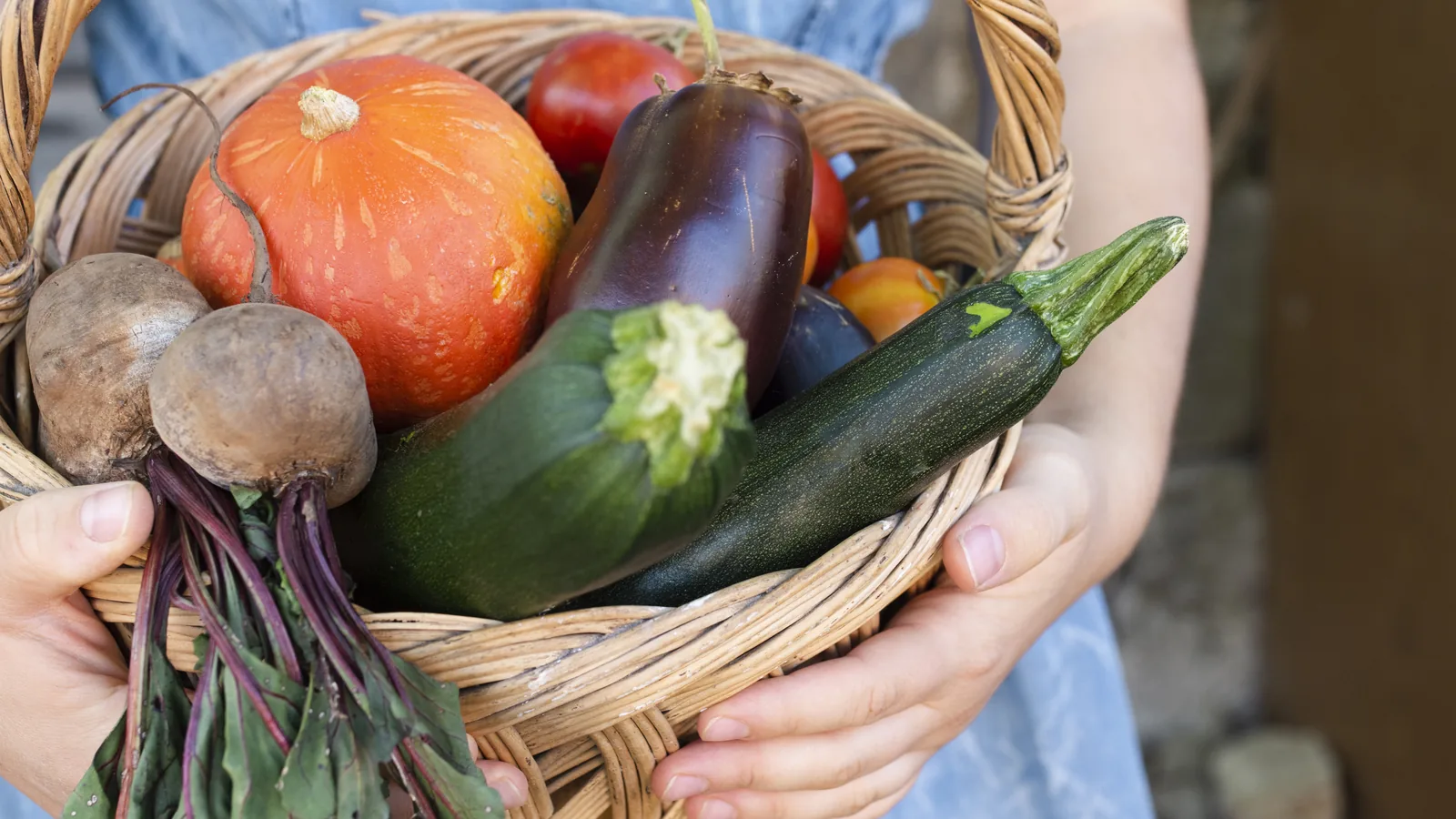 Two hands holding a wicker basket filled with freshly picked vegetables