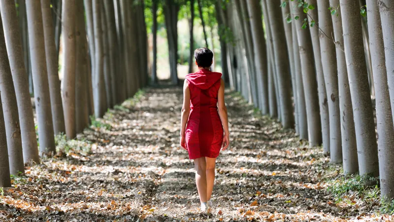 Woman in a red dress walking along a leaf-covered path between two rows of tall trees